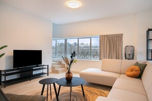 Ett modernt vardagsrum med en platt-TV, en soffa, två små bord och utsikt över en stadssiluett genom stora fönster.