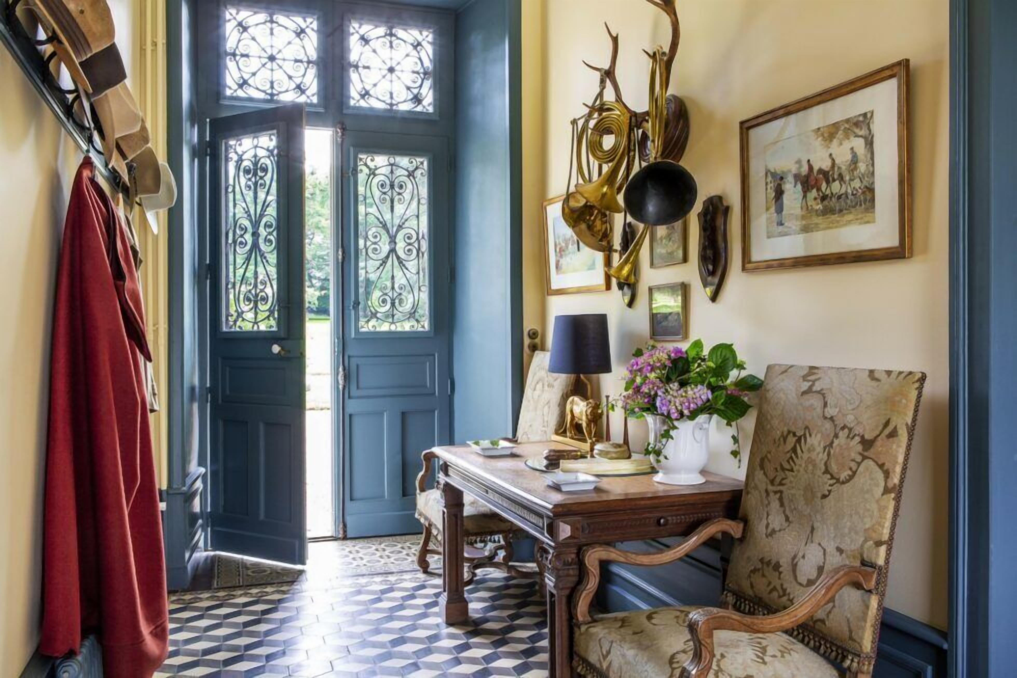 A room with a checkered floor, a blue door with decorative glass panels, a wooden table with a lamp, a chair, and wall-mounted decorations.