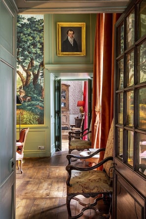A room with a wooden floor, a large window with curtains, a chair, and a painting on the wall. - Ch Teau de la Villedubois (Mordelles)