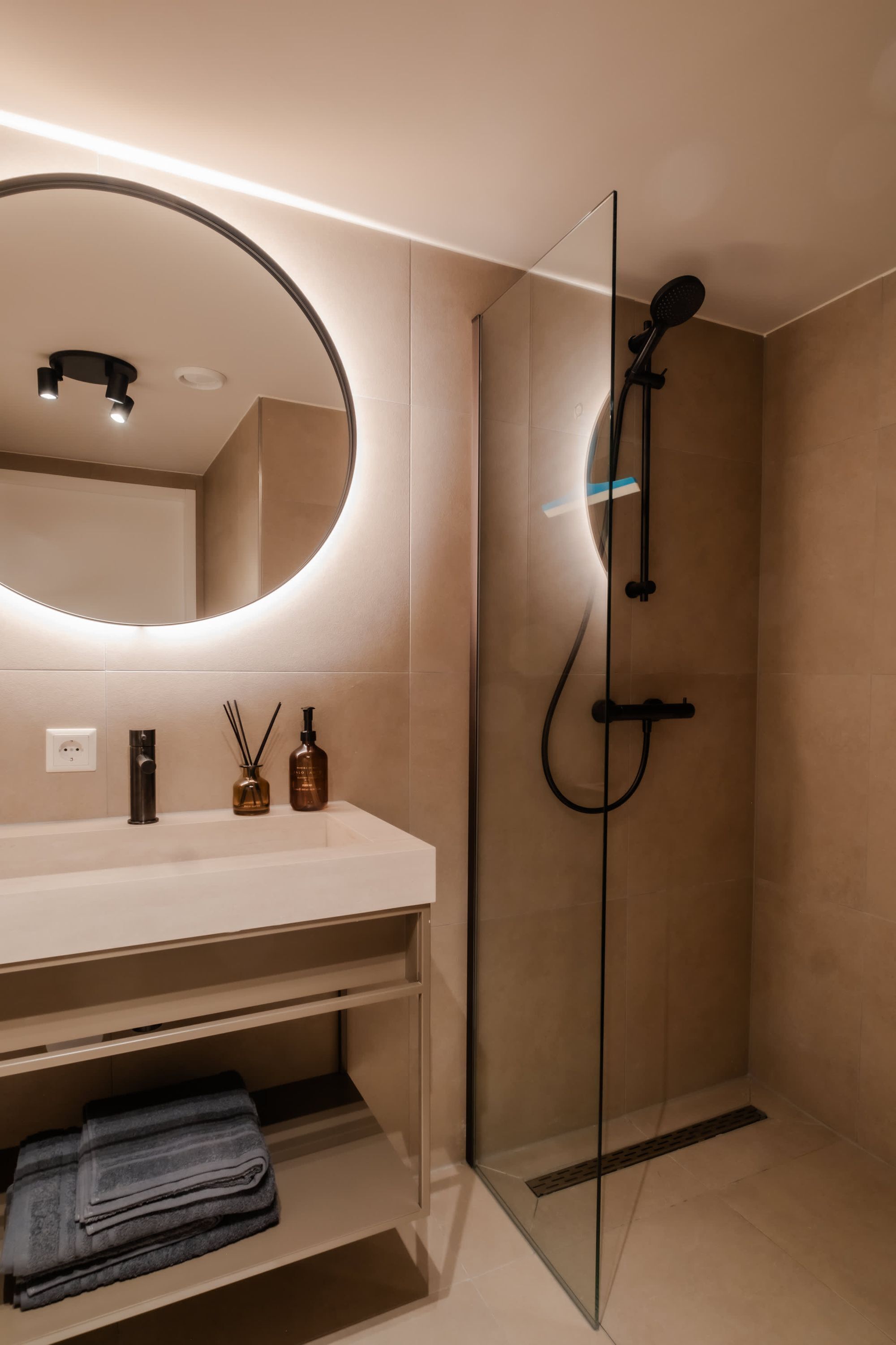 Un baño moderno con cabina de ducha de vidrio, un espejo redondo y un tocador blanco.