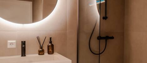 A modern bathroom with a glass-enclosed shower, a round mirror with integrated lighting, and a white vanity with a towel rack.