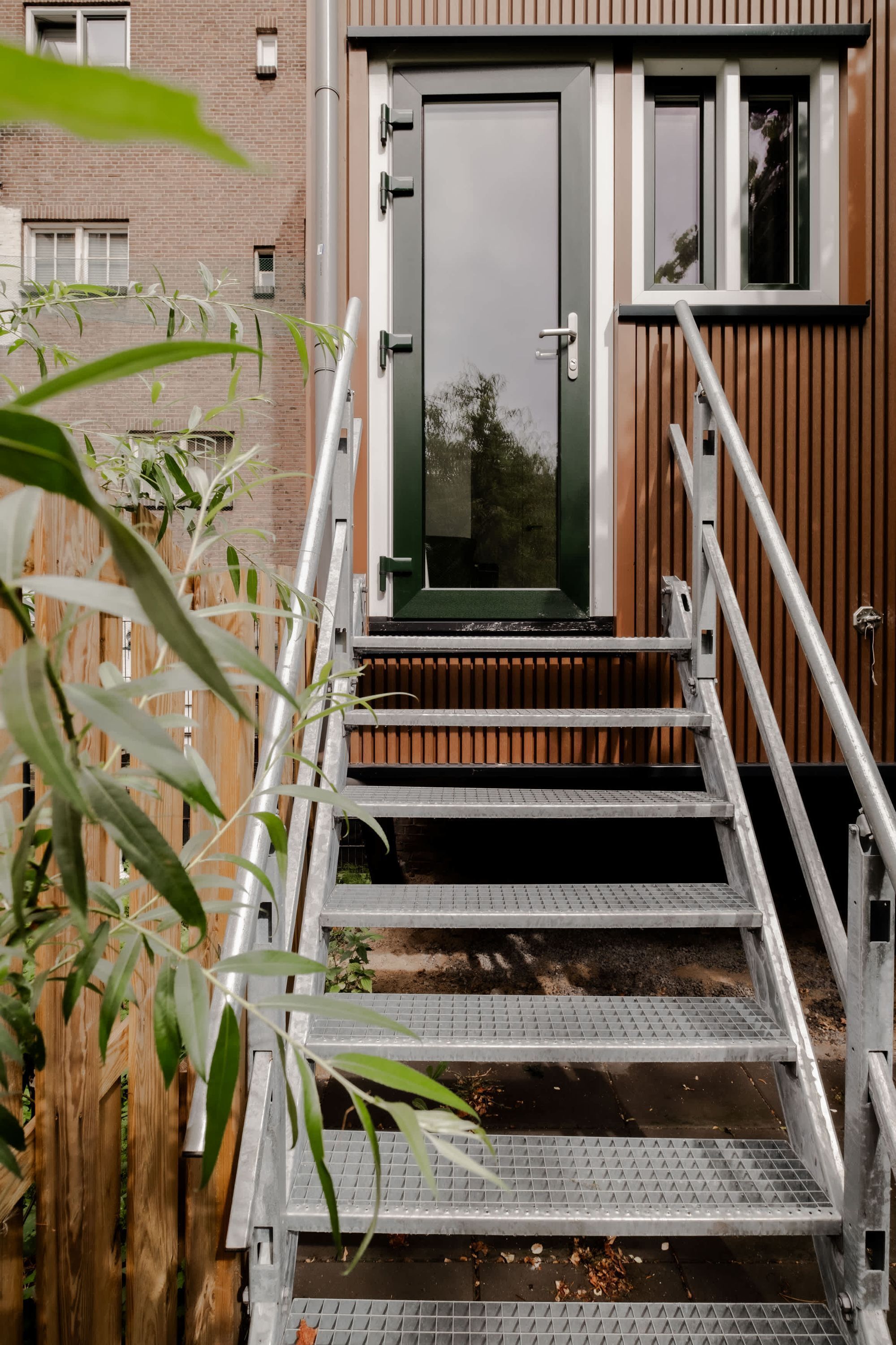 A modern building with a wooden facade, a metal staircase leading to a door, and a view of a brick building in the background.