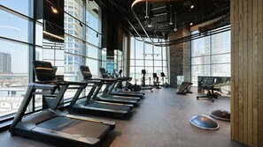 Un gimnasio con cintas de correr, bicicletas estáticas y una máquina de remo, amplias ventanas y vista a la ciudad.