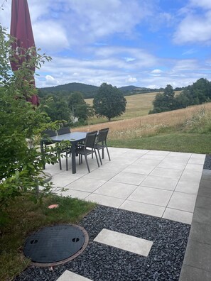 An outdoor patio with a table and chairs, surrounded by a garden and a view of the countryside.