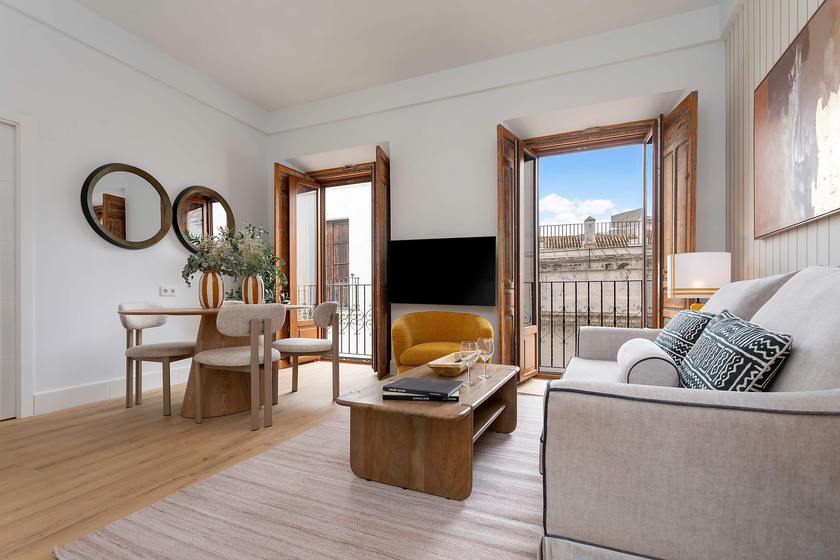Un salon moderne avec un canapé, une table basse et des fauteuils. Un téléviseur est fixé au mur et une fenêtre offre une vue sur les bâtiments.