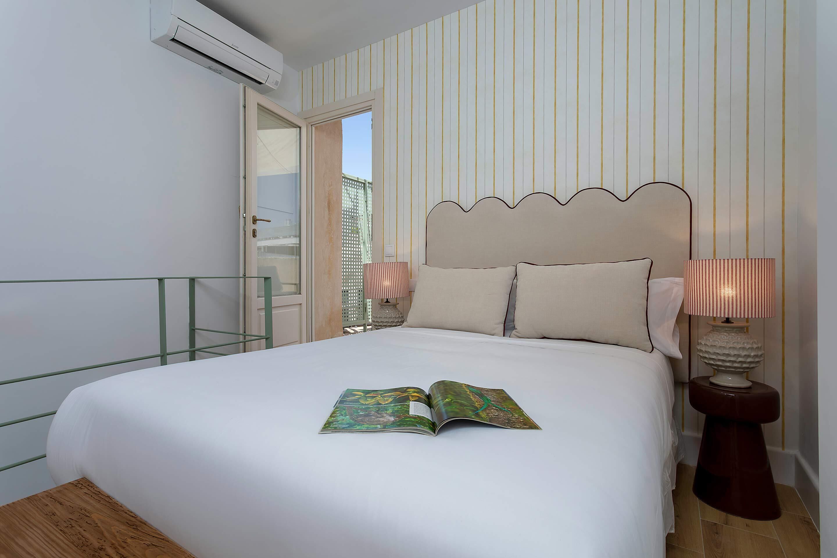 A hotel room with a bed, a nightstand with a lamp, a window with a view, and an air conditioning unit.