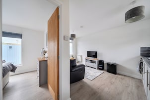 A modern living room with a wooden door, a black leather sofa, a TV, speakers, and a small kitchen area.