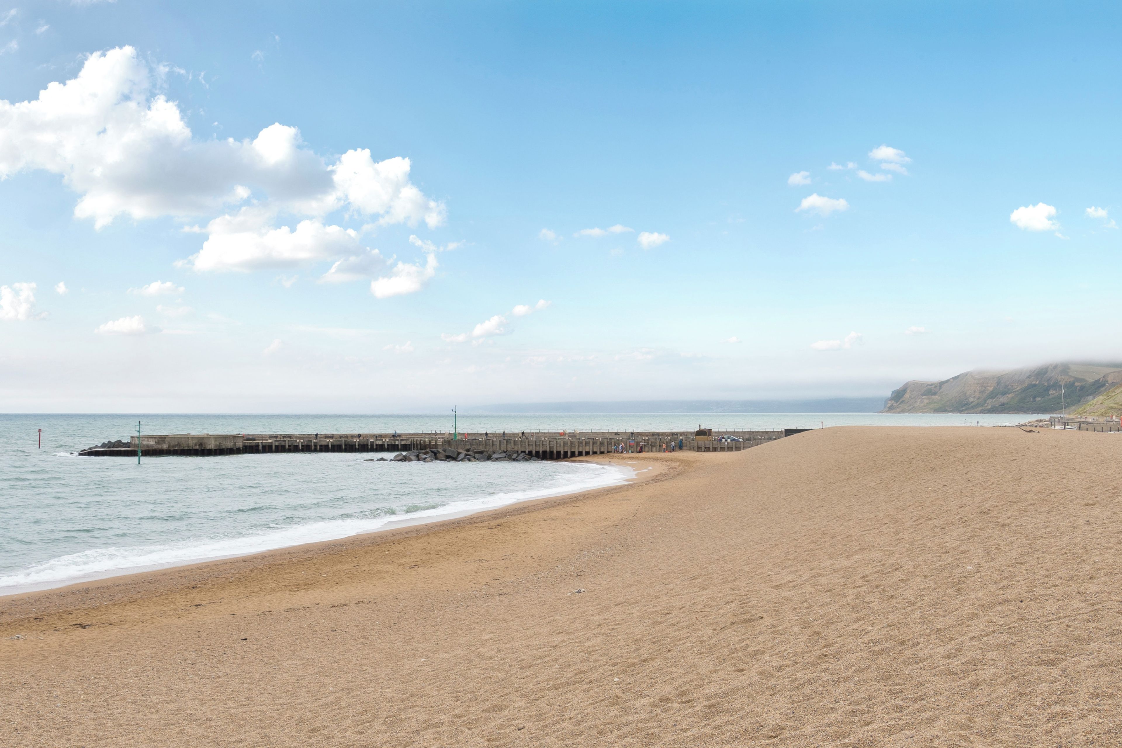 A wide sandy beach with a rocky breakwater extending into the sea, under a clear sky.