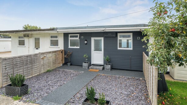 A residential building with a grey exterior, a front door, windows, and a small garden area with gravel and potted plants. - The Chalet Self Contained With Patio (Bridport)