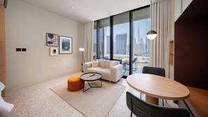 Ein modernes Hotelzimmer mit großem Fenster, einer Couch, einem runden Tisch und einem kleinen orangefarbenen Ottoman.