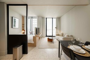 A modern hotel room with a bed, a seating area with a sofa and a coffee table, a dining table with plates and glasses, and a large window with curtains. - Furnished Business Bay Studio w Balcony (Dubai)