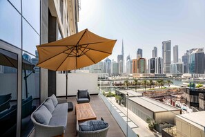 A balcony with a sofa, a table, and an umbrella, overlooking a cityscape with modern buildings. - Modern 2BR Retreat Near Downtown Dubai (Dubai)