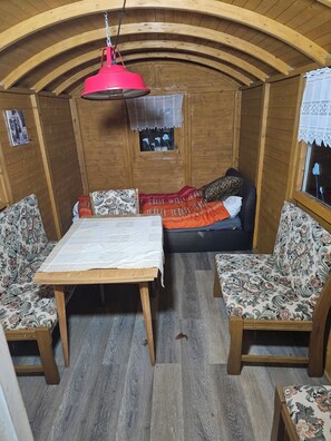 Una habitación acogedora con paredes y techo de madera, una mesa con mantel blanco, dos sillas estampadas, un sofá con una manta roja y una lámpara roja colgante.