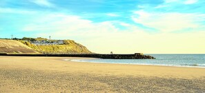 Uma praia com um molhe rochoso, um farol e um pequeno povoado numa colina.
