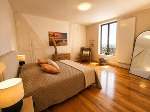Une chambre à coucher avec un lit, une chaise, une table de chevet, une lampe, une fenêtre donnant sur un beau paysage et un tableau accroché au mur.