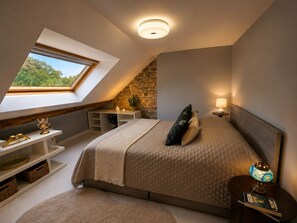Ein Schlafzimmer mit schrägen Wänden, einem Oberlicht, einer Steinwand, einem Bett mit gesteppter Bettdecke, Nachttischen und einem Beistelltisch mit Lampe.