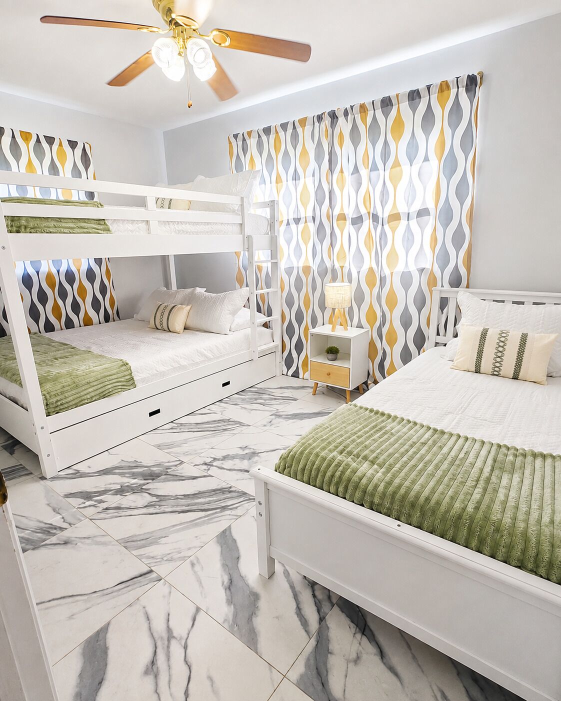 A room with a bunk bed, a single bed, a ceiling fan, a nightstand, and patterned curtains.