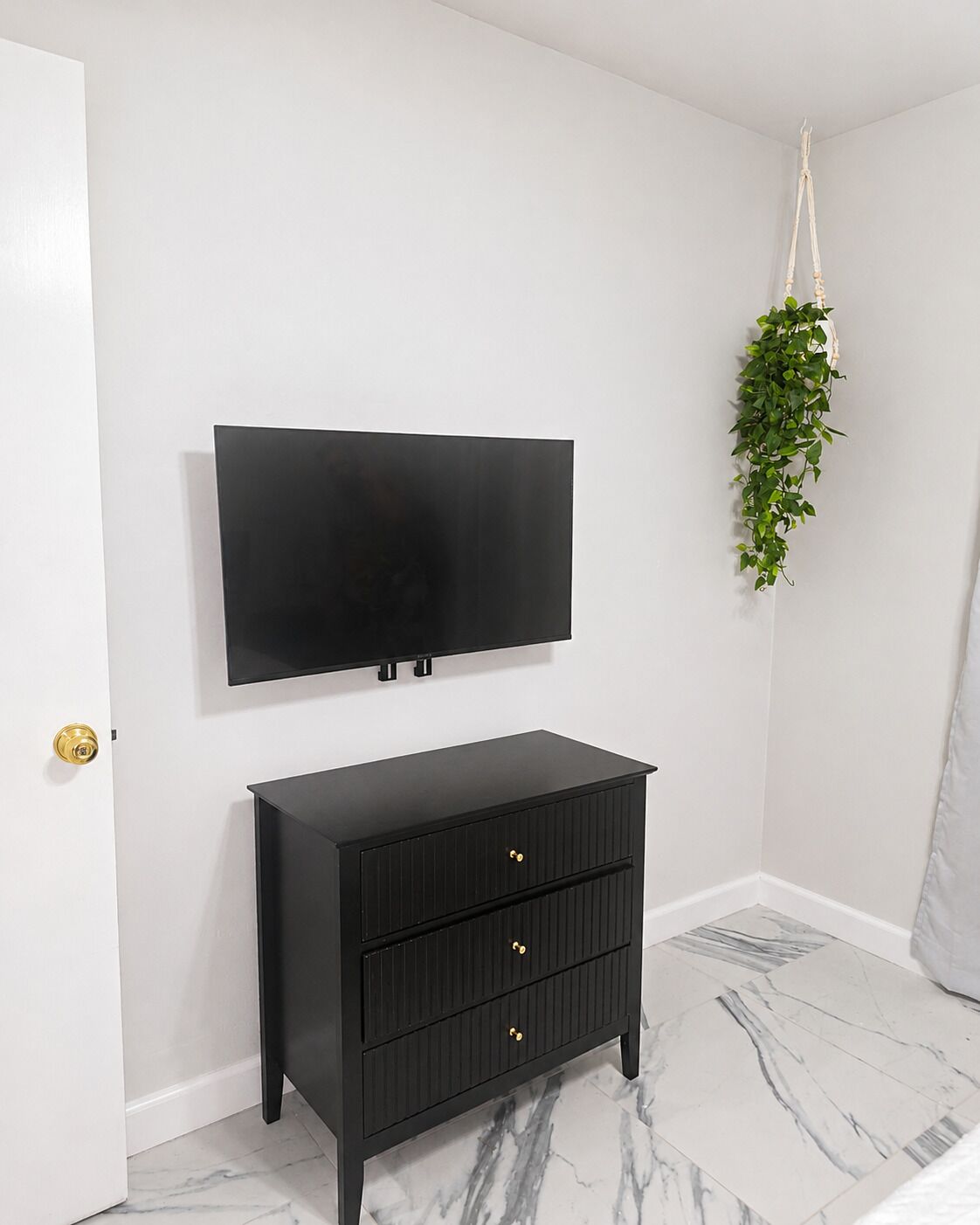 A room with a black TV mounted on the wall, a black dresser with drawers, a hanging plant, and a marble-patterned floor.