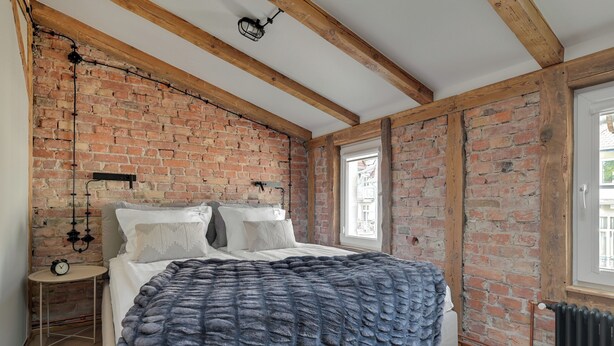 Een slaapkamer met bakstenen muren, een houten plafond, een bed met een gebreid plaid, een nachtkastje met een klok en een raam met uitzicht.