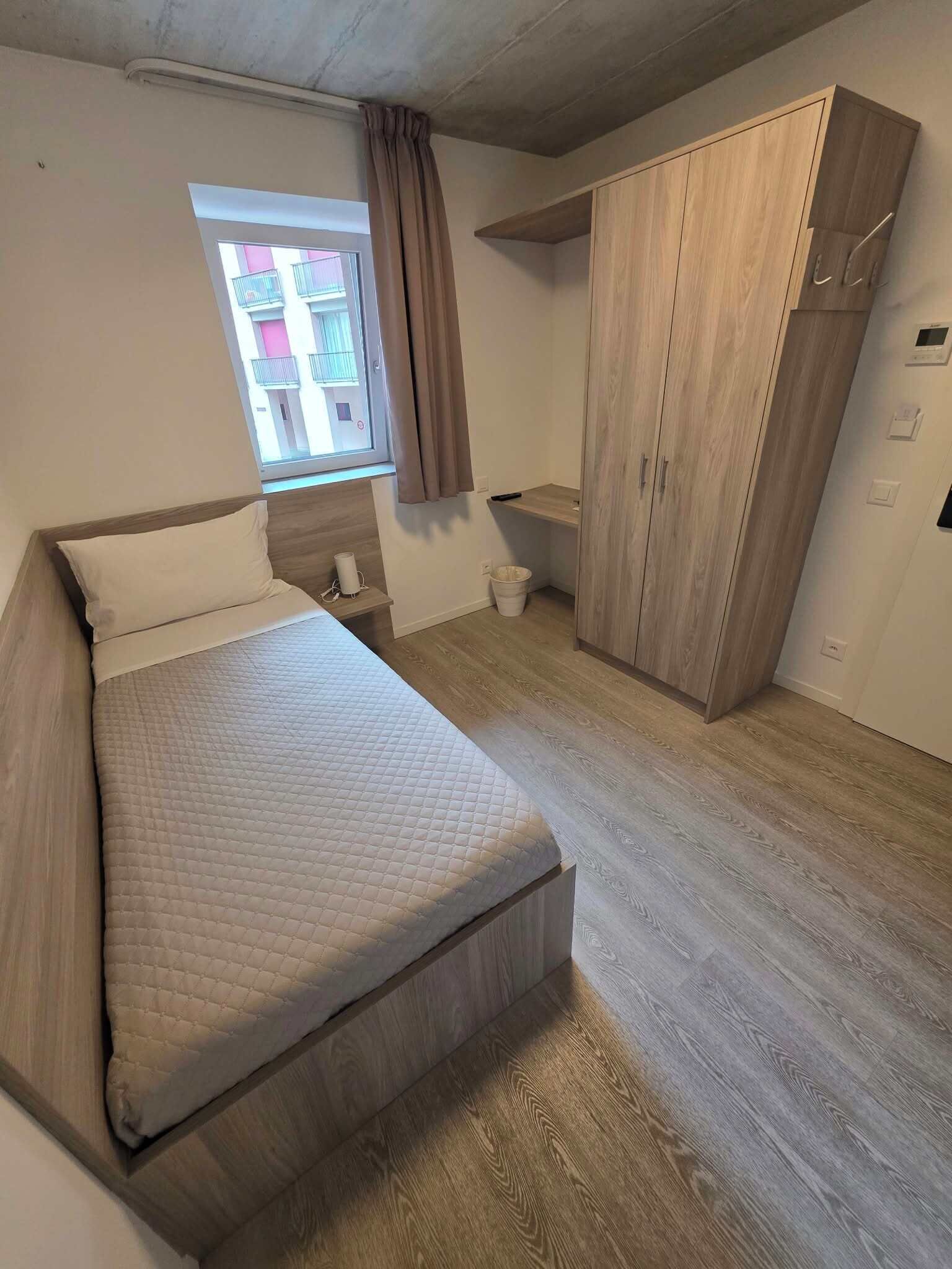 Un dormitorio con cama, armario, ventana y una mesita.