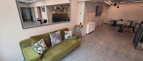Un interior moderno con un sofá verde, un espejo grande y una zona de comedor con mesas y sillas.