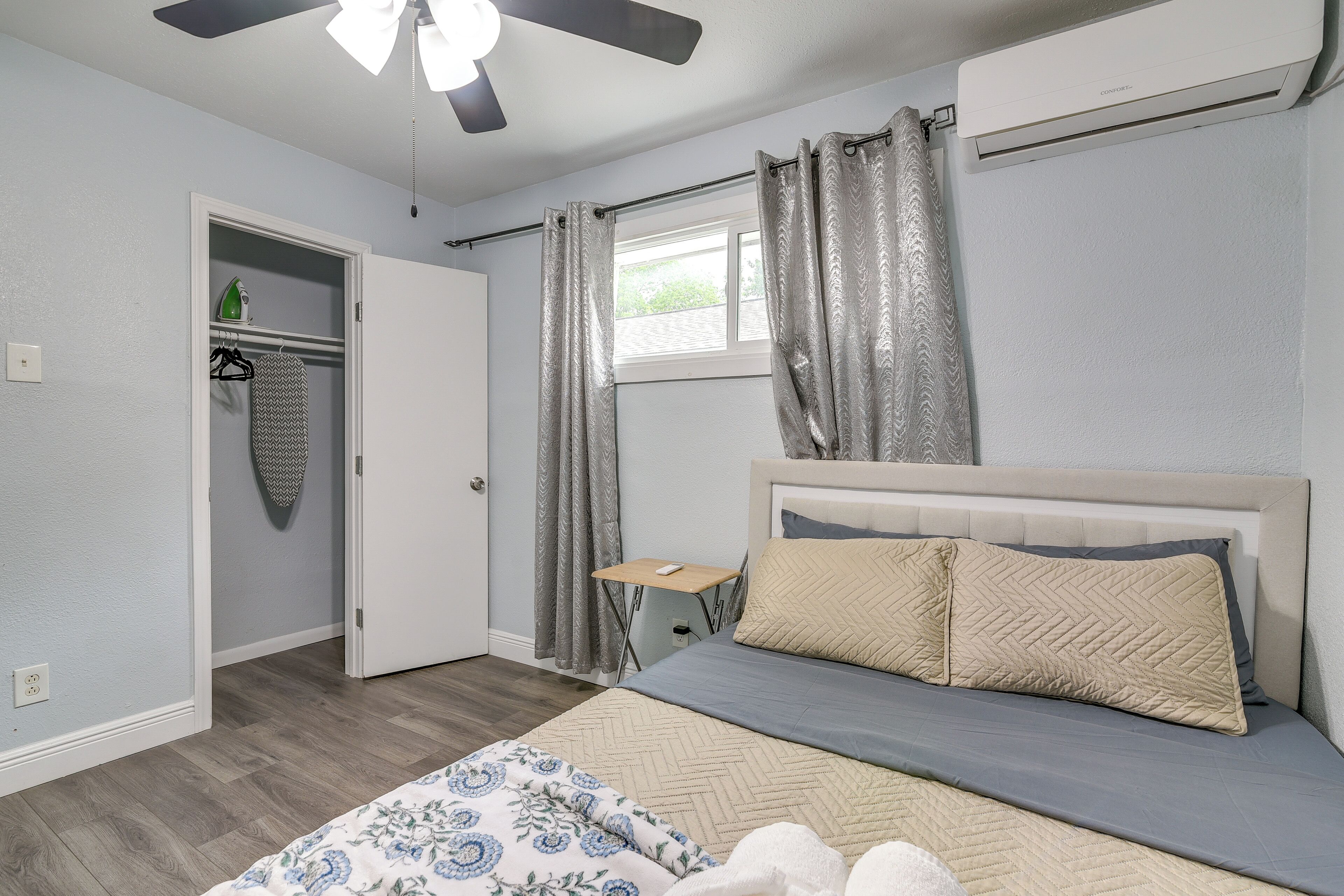 Une chambre à coucher comprenant un lit, un ventilateur de plafond, un appareil de climatisation, une fenêtre avec des rideaux, une table de chevet et une armoire.