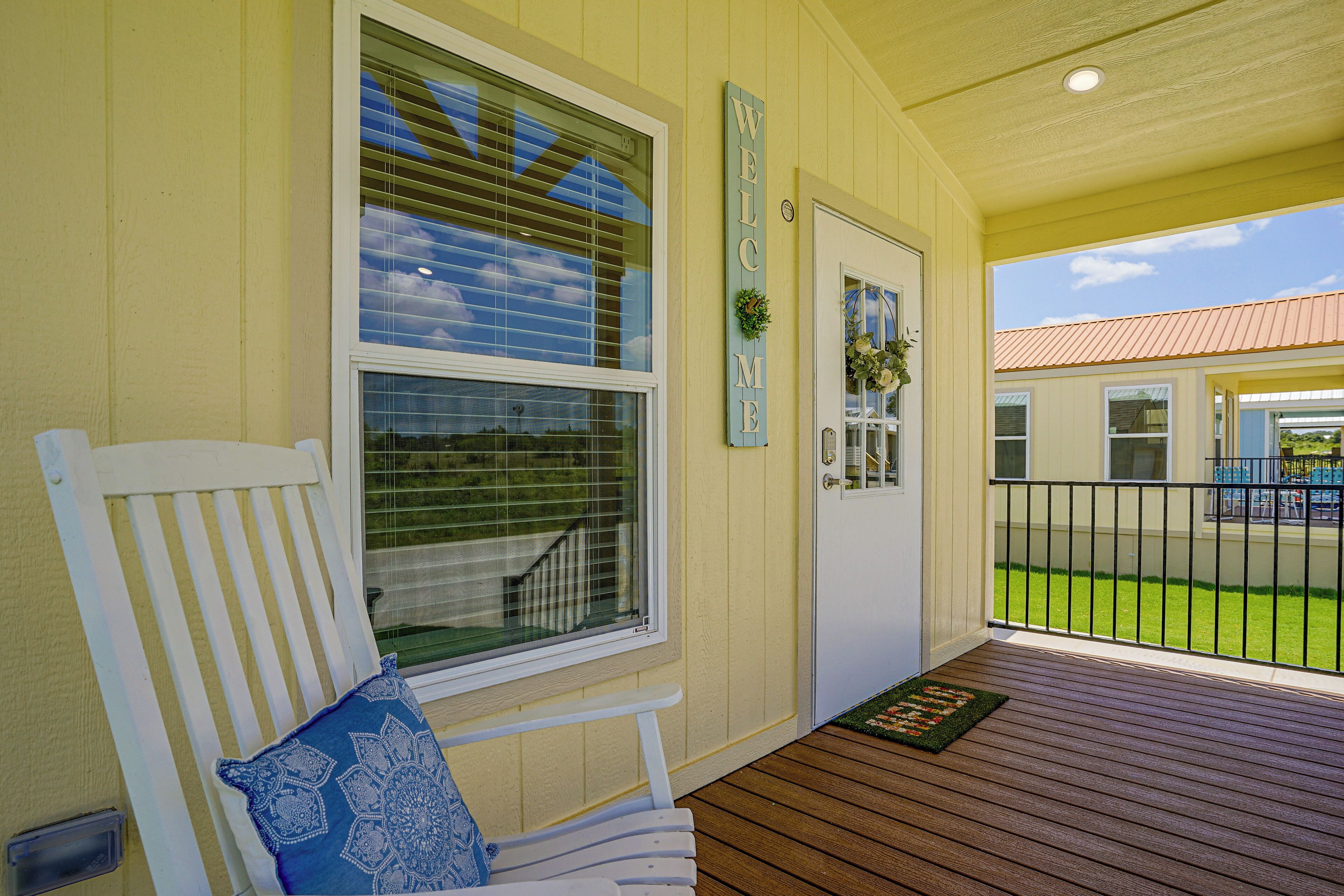 A porch with a white bench, a door with a 'WELCOME' sign, and a view of a green lawn and building across the street.