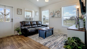 A living room with a black leather sofa, a coffee table, a ceiling fan, and large windows with blinds. - Tiny Home Retreat Close to Lake Granbury (Granbury)