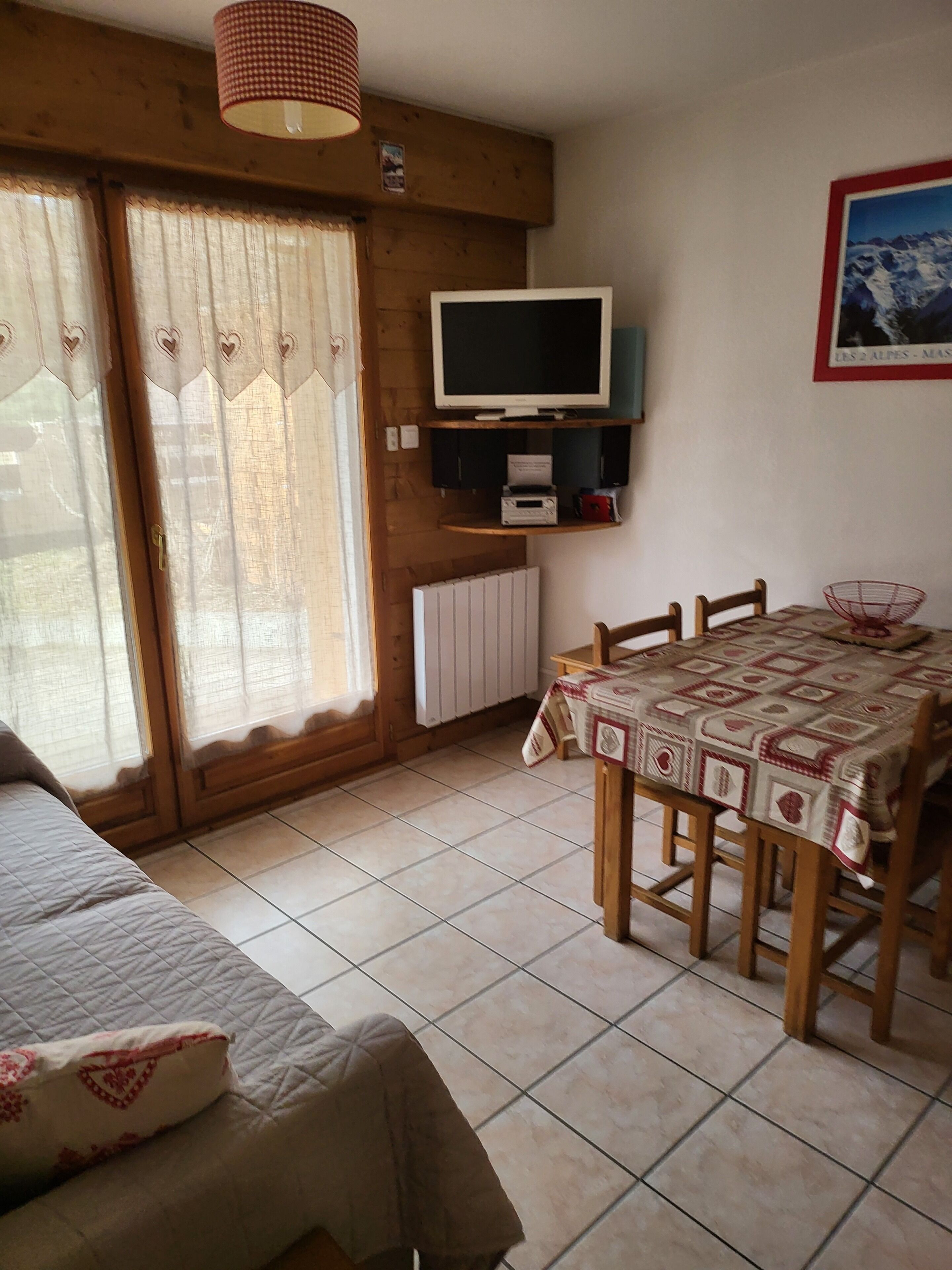 Una habitación con cama, una mesa de comedor con sillas, televisor montado en la pared y un radiador.