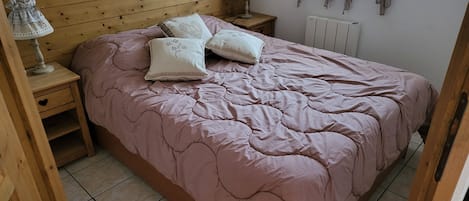 Un dormitorio con cabecera de madera, colcha rosa, dos mesitas de noche con lámparas y un radiador.