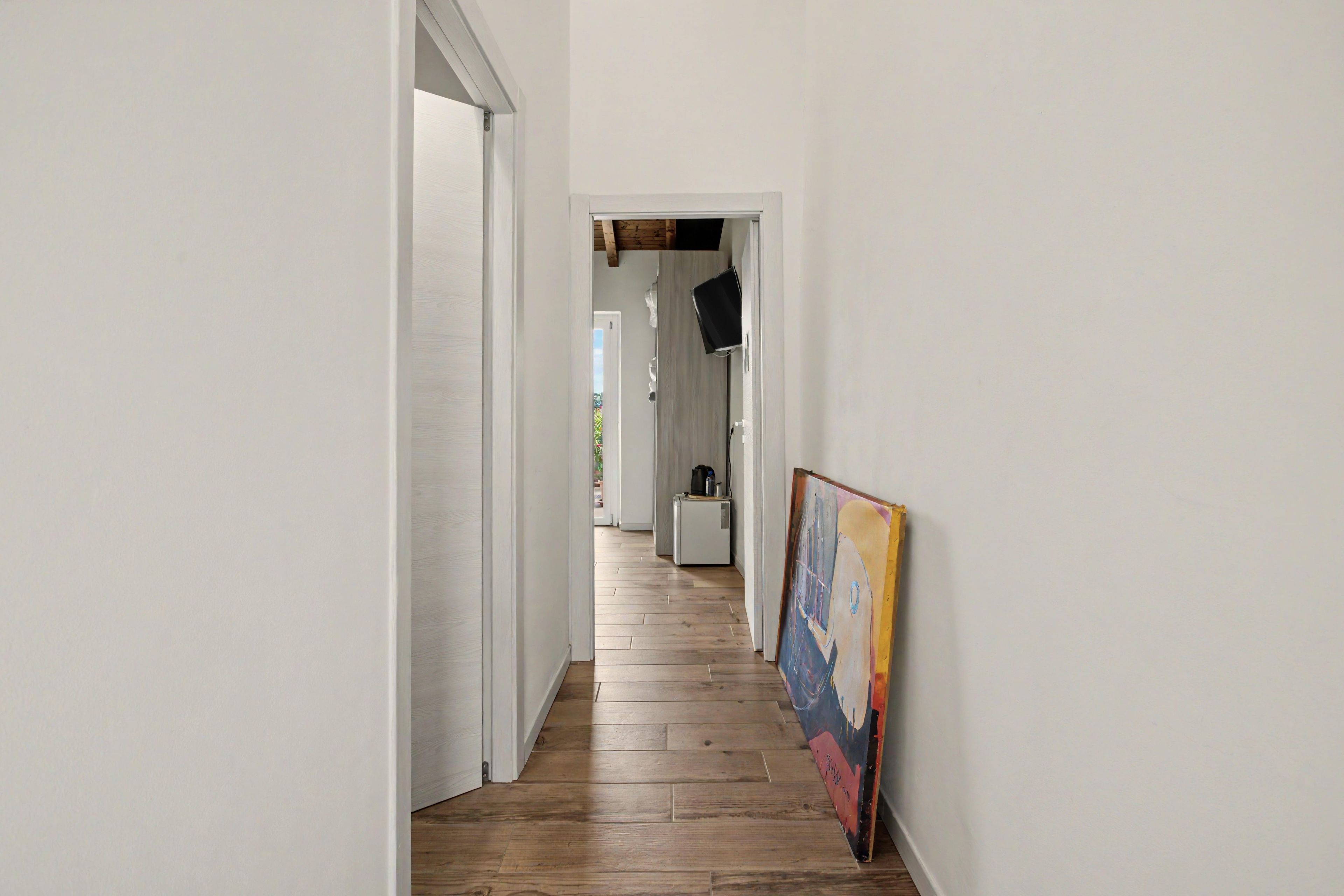 A hallway with wooden flooring, white walls, and a painting on the right.