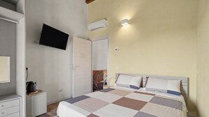 A bedroom with a bed, a television mounted on the wall, a wardrobe, and a small table with a kettle.