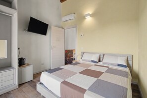 A bedroom with a bed, a television mounted on the wall, a wardrobe, and a small table with a kettle.