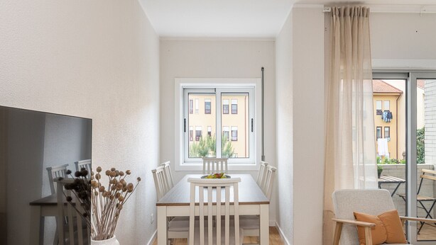 Et spiseområde med et bord og stole, et vindue med udsigt til bygninger, og en sofa med en pude.