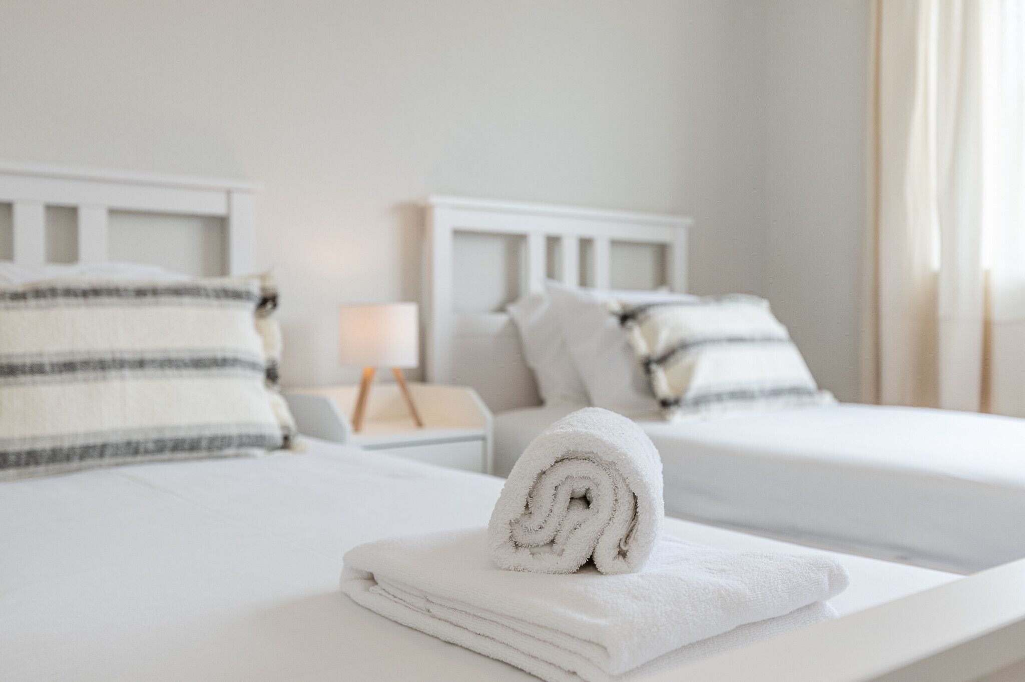 A hotel room with two beds, each with striped pillows, a neatly folded white towel, and a bedside table with a lamp.