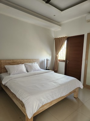 A bedroom with a wooden bed, a nightstand, a lamp, and a door. - Saren Kayutulang, Quiet 3BR Pool Villa (Badung)