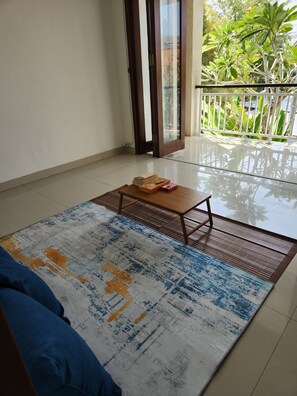 A room with a sofa, a small wooden table with books, a patterned rug, and a glass door leading to a balcony with plants. - Saren Kayutulang, Quiet 3BR Pool Villa (Badung)