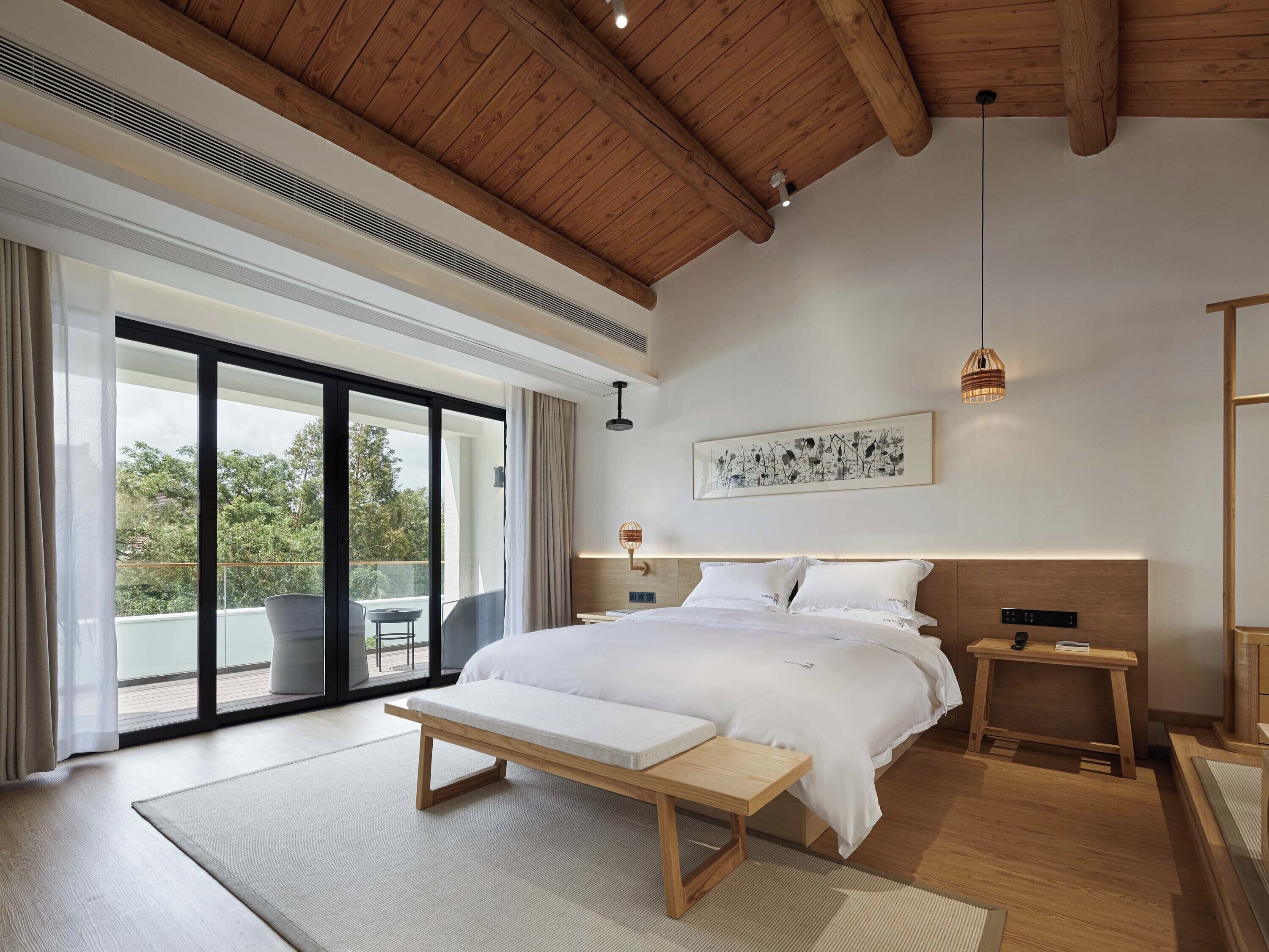 A modern bedroom with a large bed, a bench, a bedside table, and a view of the outdoors.