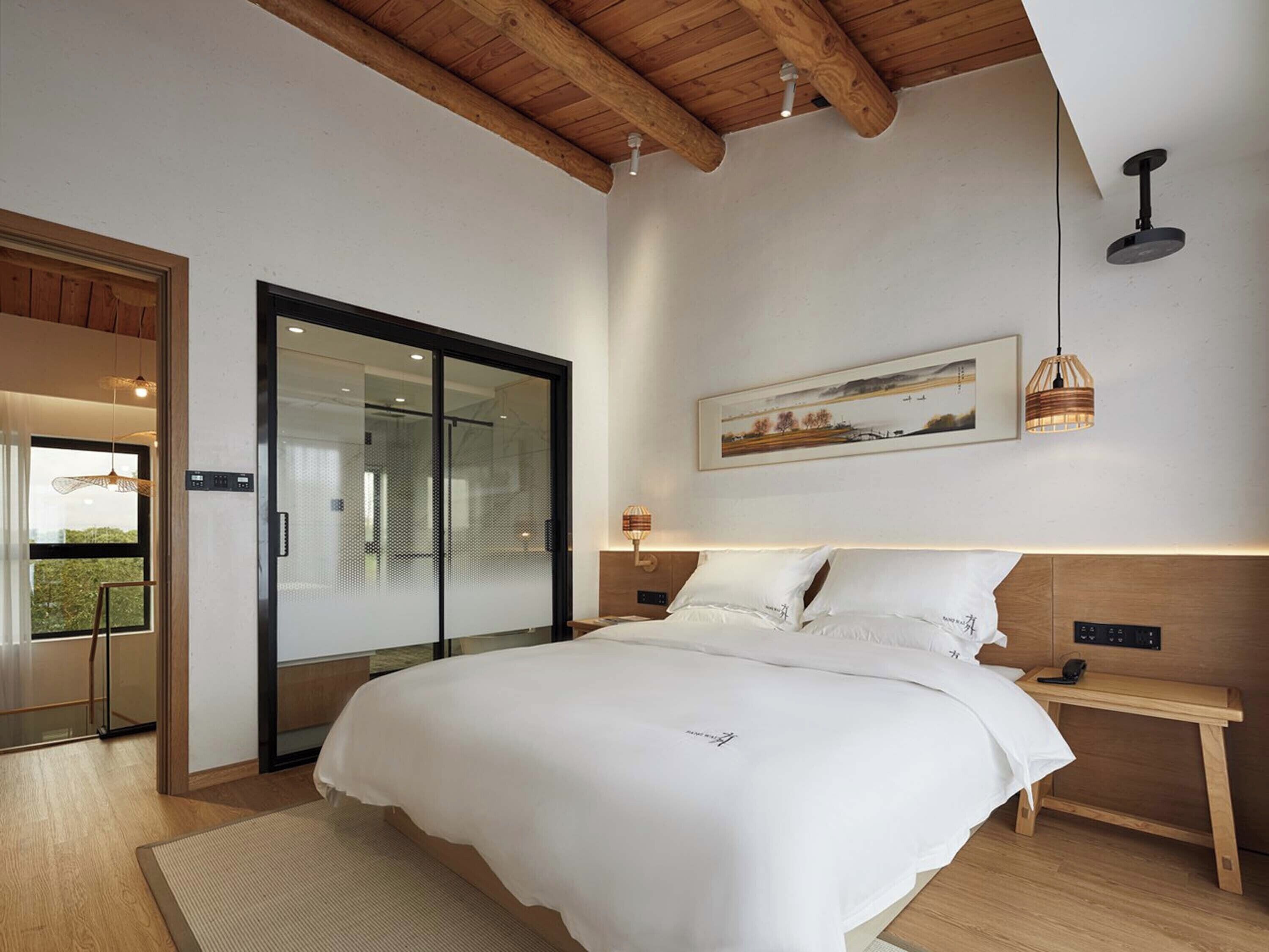 A bedroom with a large bed, a wooden headboard, a bedside table, a nightstand, a wall-mounted light fixture, and a sliding glass door leading to a balcony.