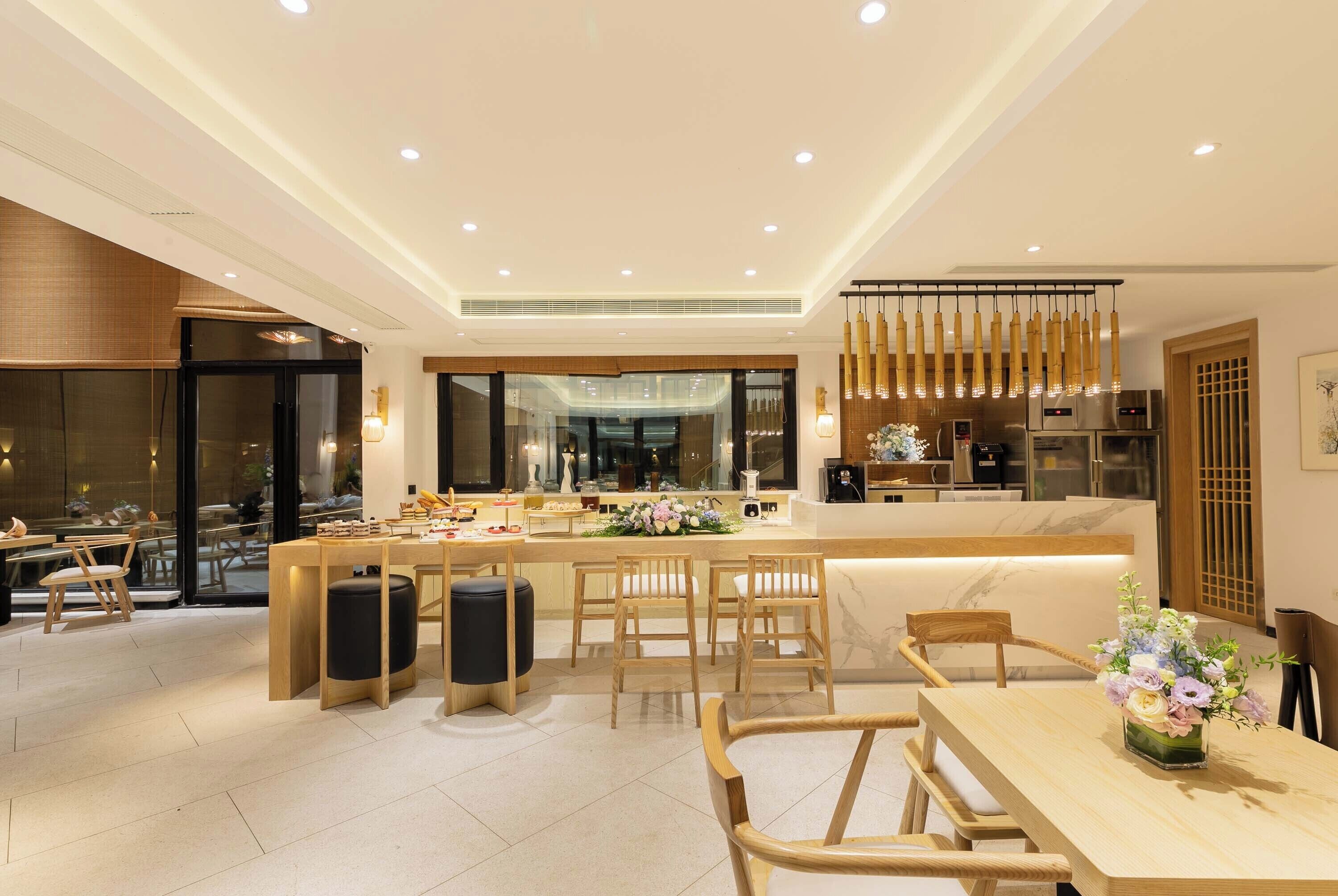 A modern hotel dining area with a central island, seating arrangements, and a kitchen visible through the glass partition.