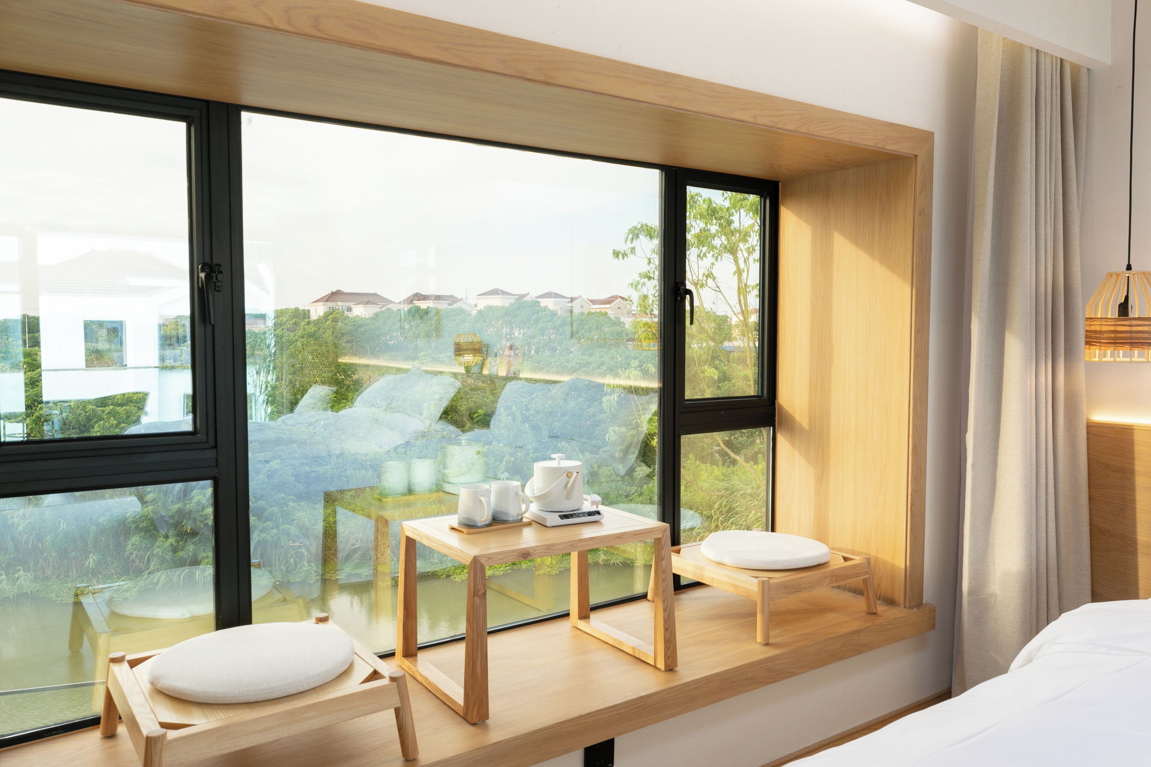 A modern hotel room with a large window, a wooden table with a kettle, cups, and a small plant.