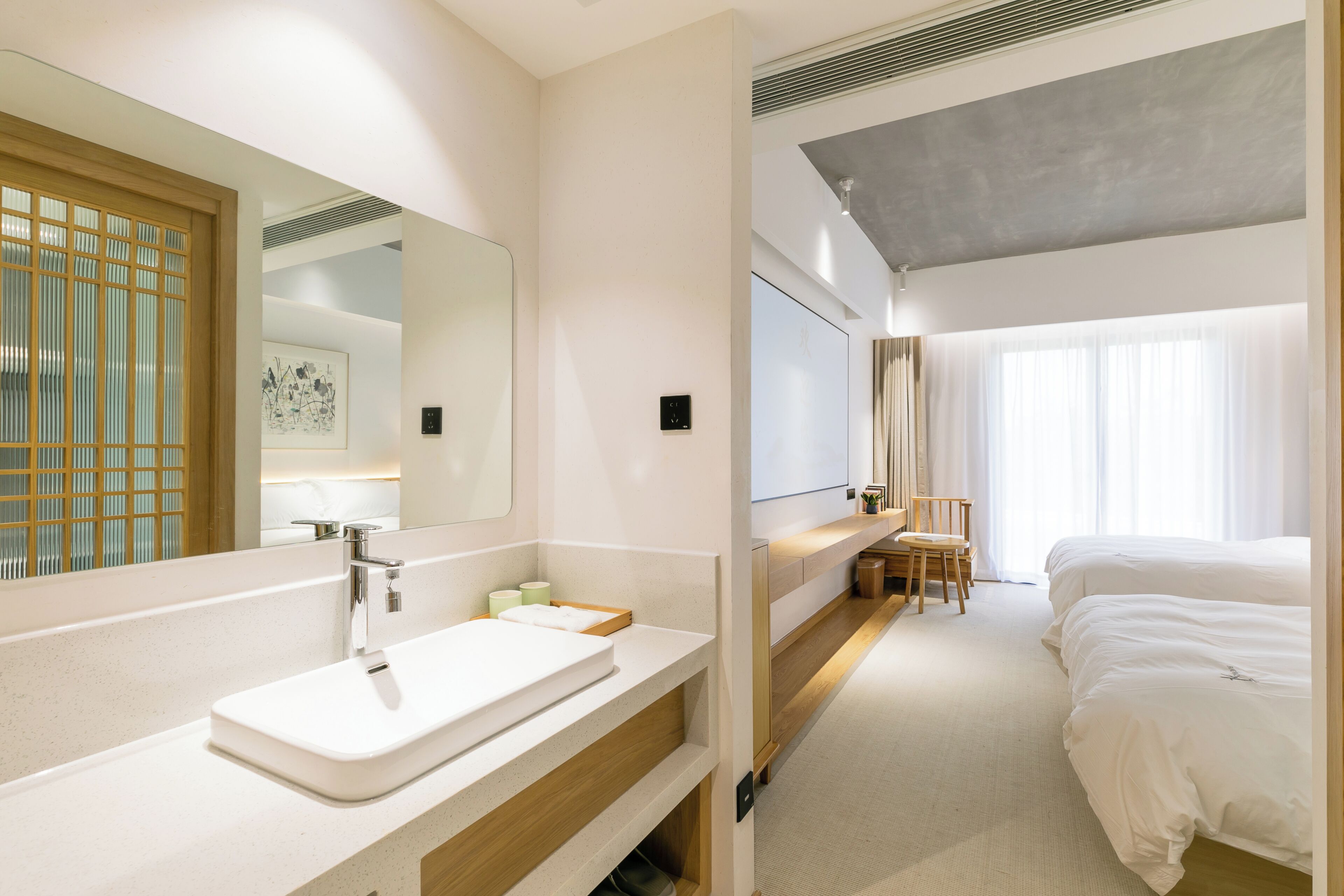 A hotel room with a large mirror, a sink, a bedside table with a lamp, a chair, and a bed.