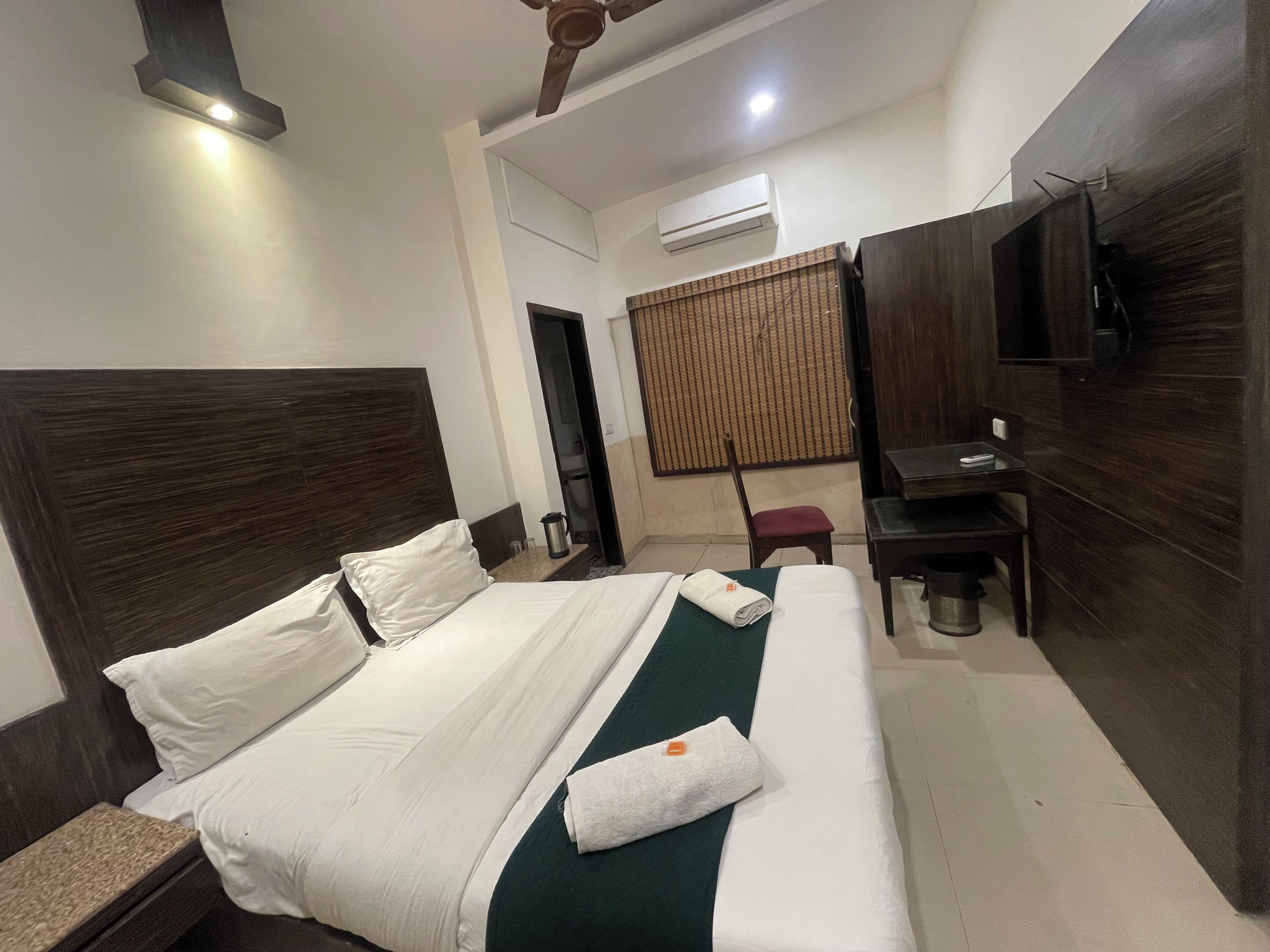 A hotel room with a bed, a desk, a chair, a TV, and an air conditioning unit.