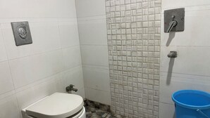 A small bathroom with a toilet, a blue bucket, and a tiled wall. - The Mohan By LIVIQ Delhi Railway Station (New Delhi)