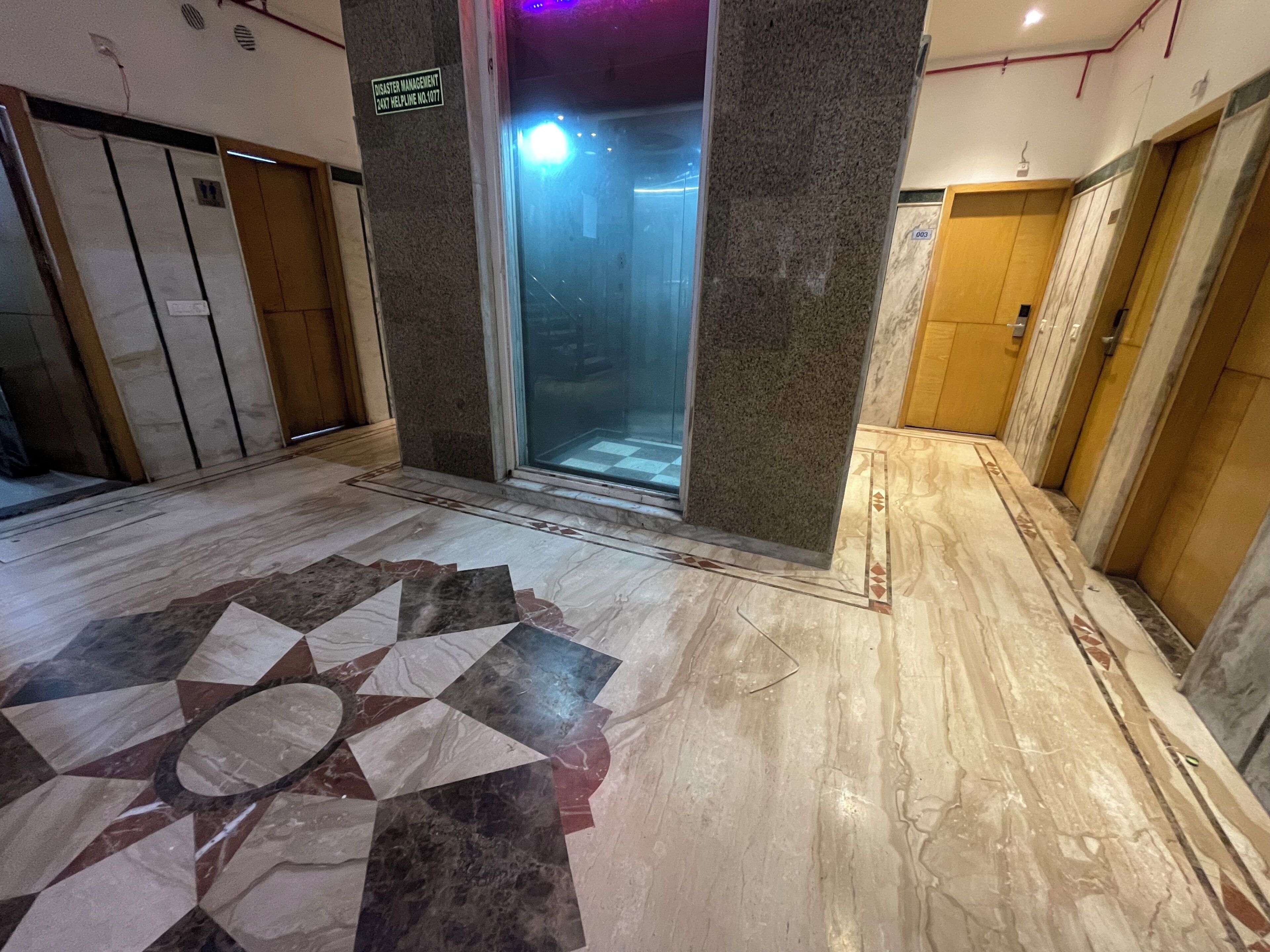 An interior corridor with a marble floor, a glass elevator door, and a sign indicating a helpline number.