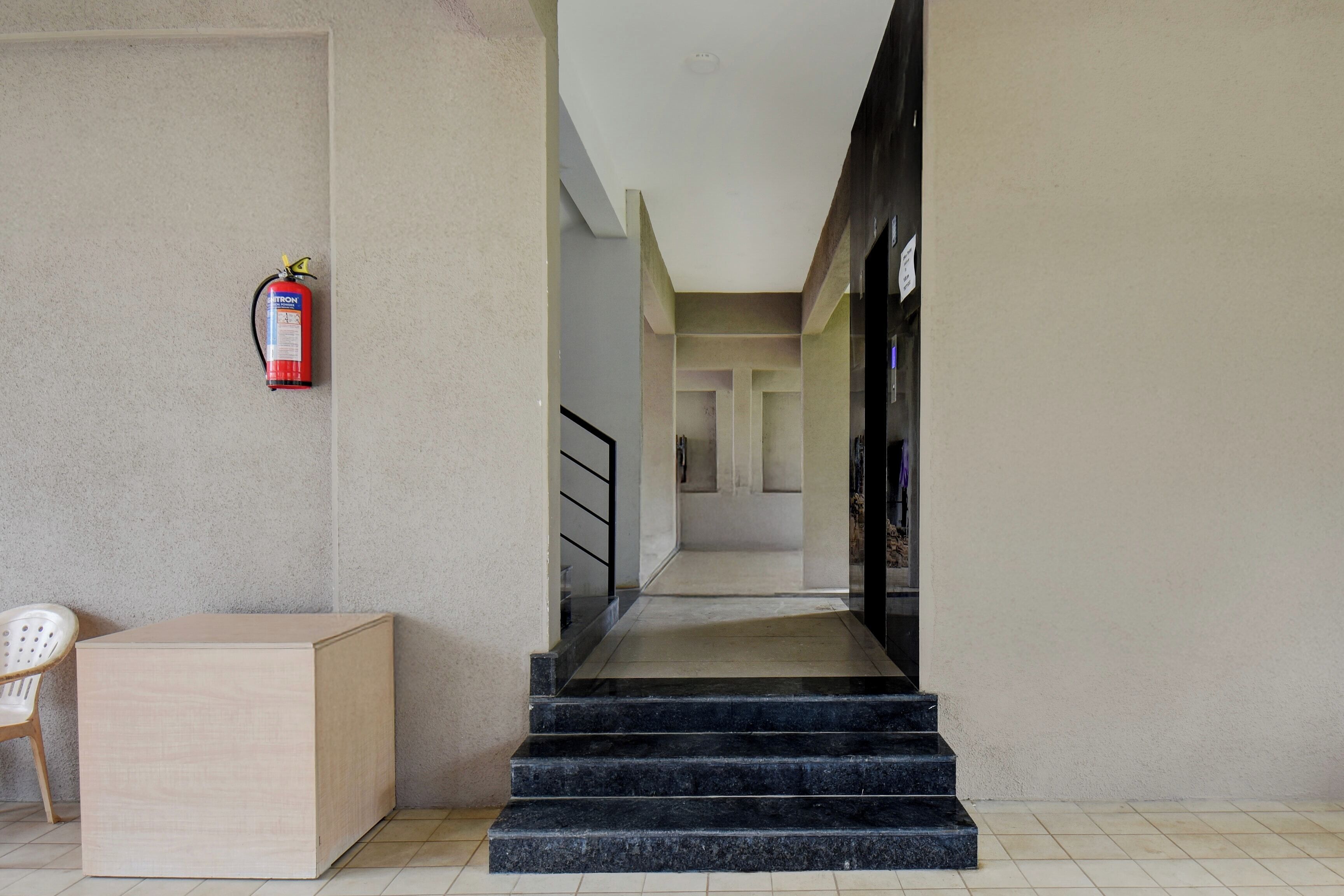 A hotel corridor with a fire extinguisher mounted on the wall, a staircase, and a chair.