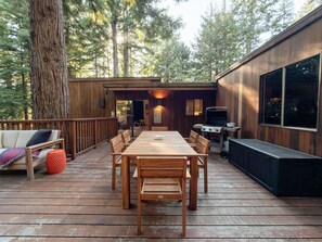 Eine Holzterrasse mit Esstisch und Stühlen, einem Grill und einem großen Fenster.