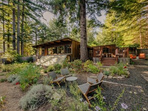 A cabin with a deck, surrounded by a garden with various plants and a seating area with chairs and a table. - Redwood Ridge | Piano, Private Sunny Deck, Hot Tub (Gualala)