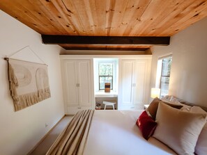Ein Schlafzimmer mit Holzboden, einem Bett mit weißer und roter Bettwäsche, einem Wandbild und einem Fenster mit Blick auf Grünflächen.