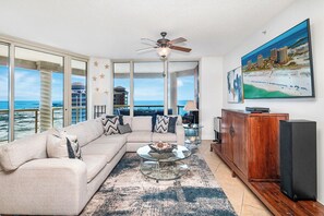 A living room with a large window, a ceiling fan, a sofa, a coffee table, and a painting of a beach scene. - 20th-Floor Portofino Sky Home with Gulf-to-Bay Views & Balcony Grill (Pensacola Beach)